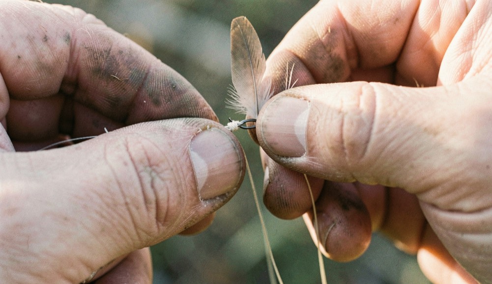 Close view of weathered fingers tying an improved clinch knot in fine tippet with a small dry fly held against soft morning light
